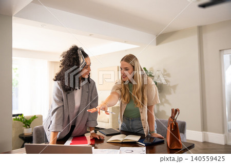Diverse female coworkers leaning over table in home office examining laptop, documents Diverse female coworkers leaning over table in home office examining laptop, documents 140559425