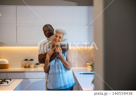 Diverse senior couple hugging in modern kitchen featuring glass jars bread box and gas cooktop 140559436