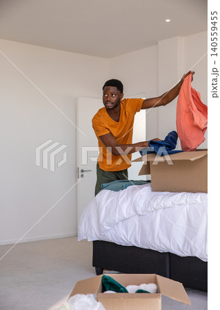 African american man sorting coral shirt, blue garments beside bed in bedroom with boxes 140559455