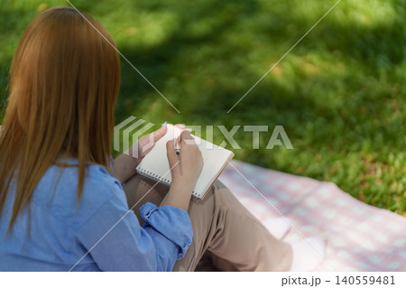Asian woman in 20s writing in notebook on picnic blanket in park Asian woman in 20s writing in notebook on picnic blanket in park 140559481
