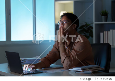 Tired Asian man yawning while working on laptop at remote home office at night Tired Asian man yawning while working on laptop at remote home office at night 140559650