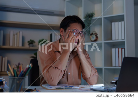 Asian man in 20s rubbing eyes from burnout working late at home office Asian man in 20s rubbing eyes from burnout working late at home office 140559664