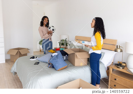 Diverse mom and teenage daughter unpacking moving boxes around bed in bedroom holding soccer ball Diverse mom and teenage daughter unpacking moving boxes around bed in bedroom holding soccer ball 140559722