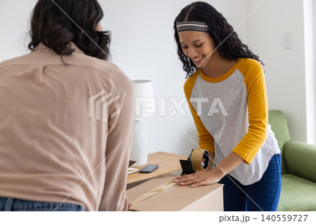 Diverse mom and teenage daughter taping cardboard box with tape dispenser on wooden table Diverse mom and teenage daughter taping cardboard box with tape dispenser on wooden table 140559727