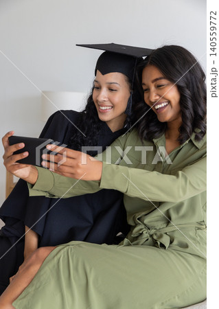 Diverse mom and teenage daughter taking selfie on sofa in graduation gown and jumpsuit Diverse mom and teenage daughter taking selfie on sofa in graduation gown and jumpsuit 140559772