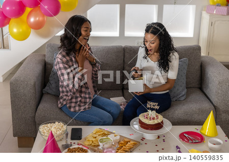 Diverse mom and teenage daughter sitting on couch, opening gift box near birthday cake and balloons 140559835