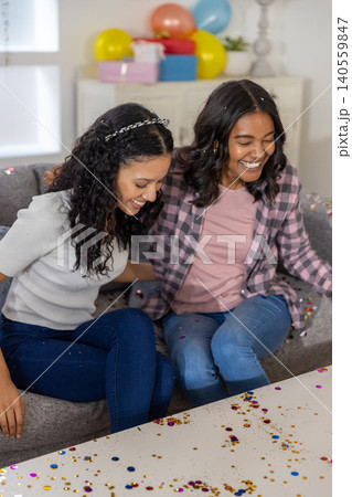 Diverse mom and teenage daughter sitting on sofa in living room with confetti, gift boxes Diverse mom and teenage daughter sitting on sofa in living room with confetti, gift boxes 140559847