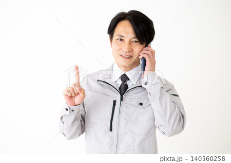 スマホで通話しながら人差し指を立てるグレーの作業着のミドル男性ビジネスマン白バック 140560258