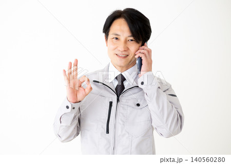 スマホで通話しながら丸のポーズをするグレーの作業着のミドル男性ビジネスマン白バック スマホで通話しながら丸のポーズをするグレーの作業着のミドル男性ビジネスマン白バック 140560280