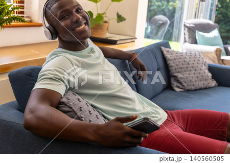 African american man reclining on sofa at home smiling holding smartphone wearing headphones 140560505