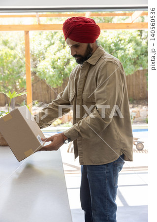 Indian man wearing red turban, shirt examining cardboard box on shaded patio table beside pool Indian man wearing red turban, shirt examining cardboard box on shaded patio table beside pool 140560536