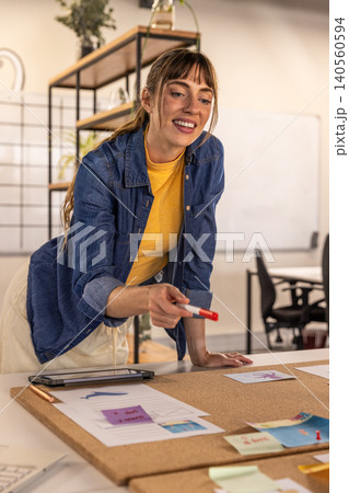 Female designer leaning over cork board in office marking sketches and sticky notes with marker 140560594