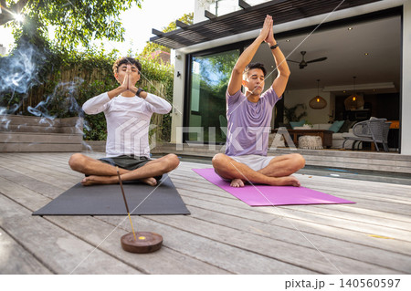 Asian male friends meditating on yoga mats on wooden deck at home with incense stick 140560597