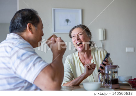 Diverse couple sipping coffee at dining table with french press, laughing with smartphone 140560638