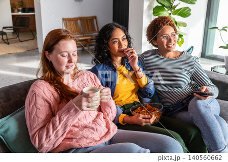 Diverse female friends watching show on sofa at home, holding mug and pretzels, with plants 140560662