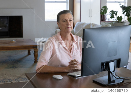 Senior woman wearing shirt working at living room desk using widescreen monitor and keyboard Senior woman wearing shirt working at living room desk using widescreen monitor and keyboard 140560750