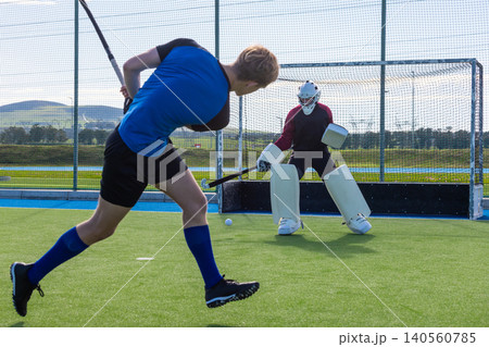 Male opponents swinging hockey stick and striking ball at goalkeeper on hockey pitch, copy space Male opponents swinging hockey stick and striking ball at goalkeeper on hockey pitch, copy space 140560785