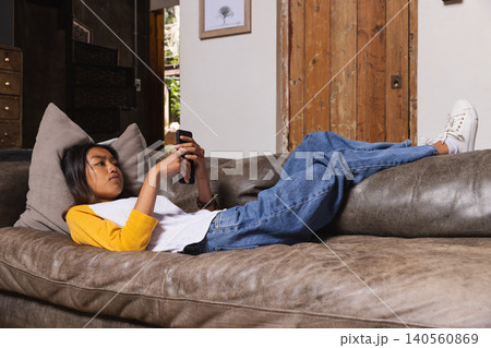 Teenage Asian female reclining on brown sofa with grey pillow in living room, holding smartphone 140560869