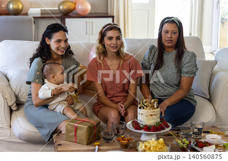 Diverse mothers with baby and friend sitting on couch celebrating birthday with cake and gift box 140560875