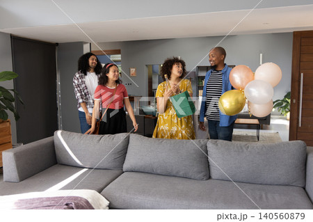 Diverse friends standing by grey sofa entering living room with green gift bag, balloon cluster Diverse friends standing by grey sofa entering living room with green gift bag, balloon cluster 140560879