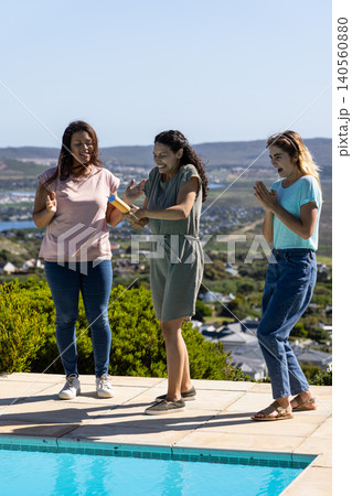 Diverse female friends spraying with water gun on tiled pool deck by pool overlooking coast Diverse female friends spraying with water gun on tiled pool deck by pool overlooking coast 140560880