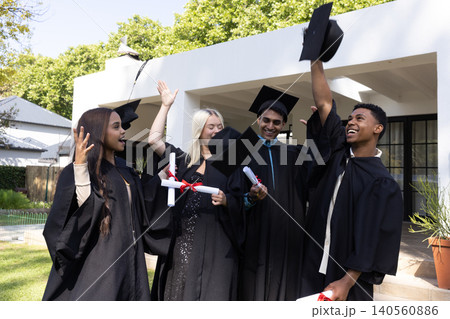 Diverse classmates celebrating on campus lawn wearing black gowns tossing caps, holding diplomas Diverse classmates celebrating on campus lawn wearing black gowns tossing caps, holding diplomas 140560886