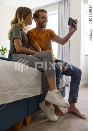 Couple smiling and taking selfie on bed edge in bedroom with smartphone and green potted plant Couple smiling and taking selfie on bed edge in bedroom with smartphone and green potted plant 140560936
