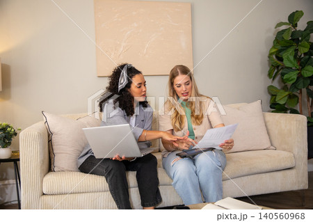 Female colleagues reviewing documents on beige sofa in living room with silver laptop, notebook Female colleagues reviewing documents on beige sofa in living room with silver laptop, notebook 140560968