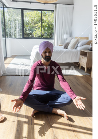 Indian man wearing lavender turban sitting meditating on bedroom floor by large window, bed Indian man wearing lavender turban sitting meditating on bedroom floor by large window, bed 140561118