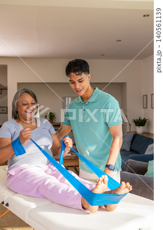 Senior african american patient, hispanic therapist working with band on table at home Senior african american patient, hispanic therapist working with band on table at home 140561139