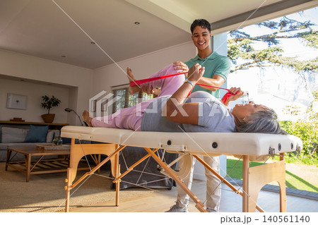 Senior female patient lying on therapy table and therapist stretching leg with band 140561140