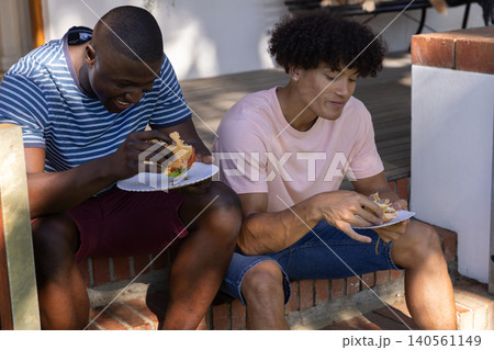 Diverse friends sitting on brick front porch steps eating hamburgers from plates in sunlight Diverse friends sitting on brick front porch steps eating hamburgers from plates in sunlight 140561149
