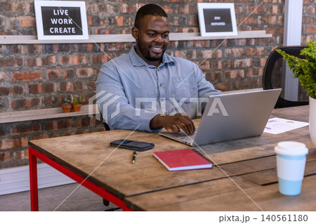 African american man typing on laptop at desk in workspace with coffee, plants, framed prints African american man typing on laptop at desk in workspace with coffee, plants, framed prints 140561180