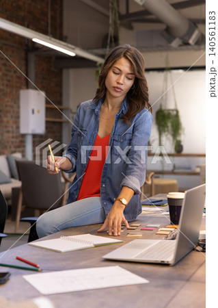 Female designer sitting on desk in modern coworking office reviewing color swatches with laptop Female designer sitting on desk in modern coworking office reviewing color swatches with laptop 140561183