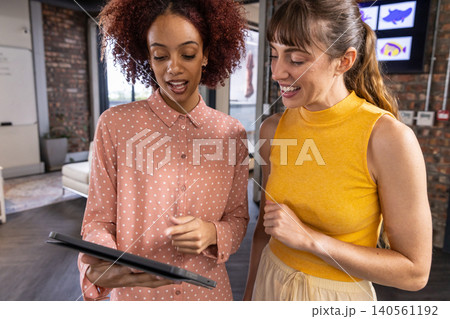 Diverse female coworkers studying tablet screen in office with brick wall fish graphics Diverse female coworkers studying tablet screen in office with brick wall fish graphics 140561192