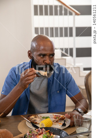 Mature adult African American man eating grilled chicken wing with corn cob at home dining table Mature adult African American man eating grilled chicken wing with corn cob at home dining table 140561211