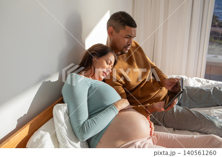 Diverse female couple expecting baby and leaning back on pillows while scrolling tablet in bedroom Diverse female couple expecting baby and leaning back on pillows while scrolling tablet in bedroom 140561260