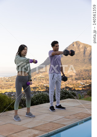 Diverse friends lifting dumbbells on stone patio near swimming pool under clear sky 140561359