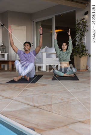 Diverse couple practicing yoga sitting cross-legged raising arms overhead at poolside on mats Diverse couple practicing yoga sitting cross-legged raising arms overhead at poolside on mats 140561361