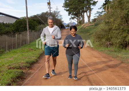 Senior indian couple jogging together on dirt road wearing running shoes, passing wire fence 140561373