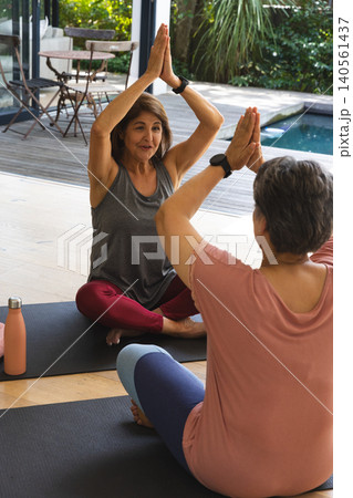 Diverse senior female friends stretching on black yoga mats near metal bistro table by pool deck Diverse senior female friends stretching on black yoga mats near metal bistro table by pool deck 140561437