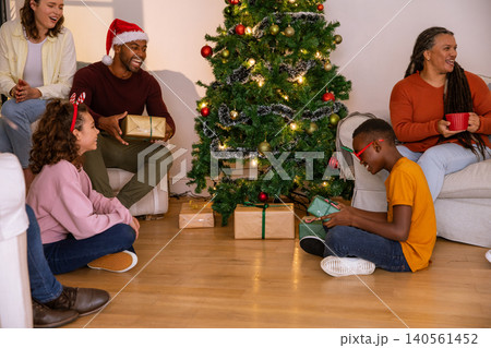 Diverse family unwrapping gifts, christmas tree with string lights in living room Diverse family unwrapping gifts, christmas tree with string lights in living room 140561452