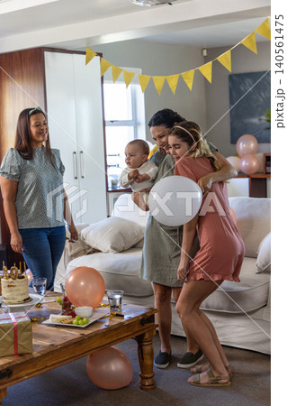 Diverse female friends holding baby boy and hugging in living room with cake, gift, balloons 140561475
