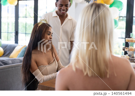 Diverse friends celebrating birthday around table at home with balloons, tiara, corset dress 140561494