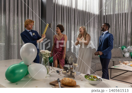 Diverse wedding party in formal attire releasing gold confetti with popper around living room Diverse wedding party in formal attire releasing gold confetti with popper around living room 140561513