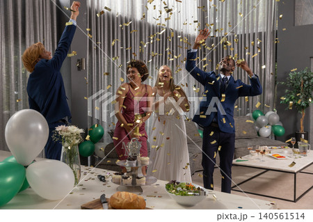 Diverse friends celebrating under falling gold confetti in living room with cupcakes, trophy 140561514