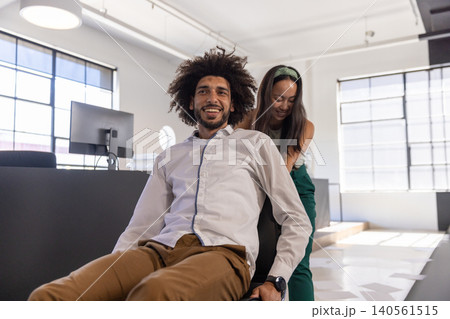 Diverse coworkers pushing rolling office chair with smiling man at office near monitor 140561515