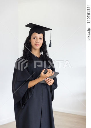 Teenage female student in graduation gown and mortarboard holding tablet device in plain room Teenage female student in graduation gown and mortarboard holding tablet device in plain room 140561524