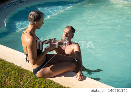 Senior couple offering water glasses sitting at backyard pool edge with grass, copy space Senior couple offering water glasses sitting at backyard pool edge with grass, copy space 140561542