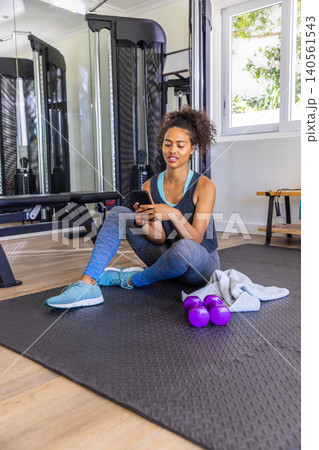 African American adult female sitting on mat in home gym holding smartphone, dumbbells nearby African American adult female sitting on mat in home gym holding smartphone, dumbbells nearby 140561543